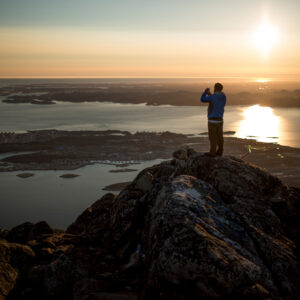 Person standing on a rocky peak, taking a photo of the sunset over the water