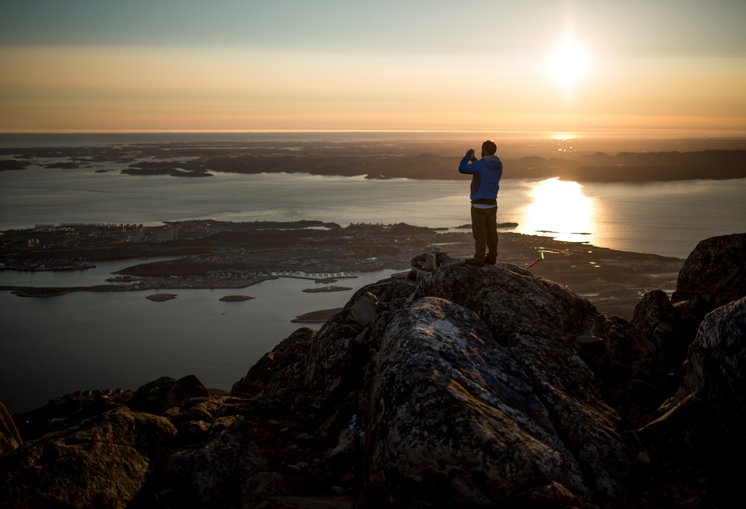 Person standing on a rocky peak, taking a photo of the sunset over the water