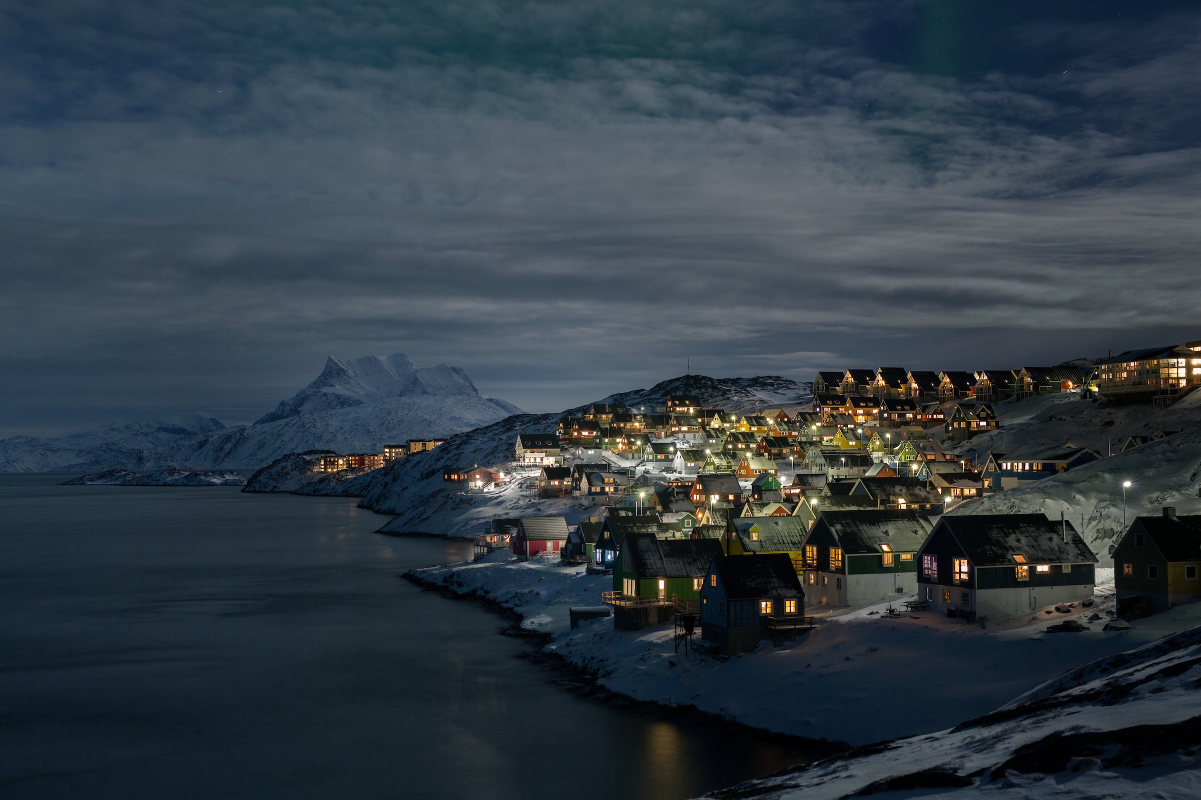 Snowy coastal village lit at night beside a calm fjord, with mountains in the background