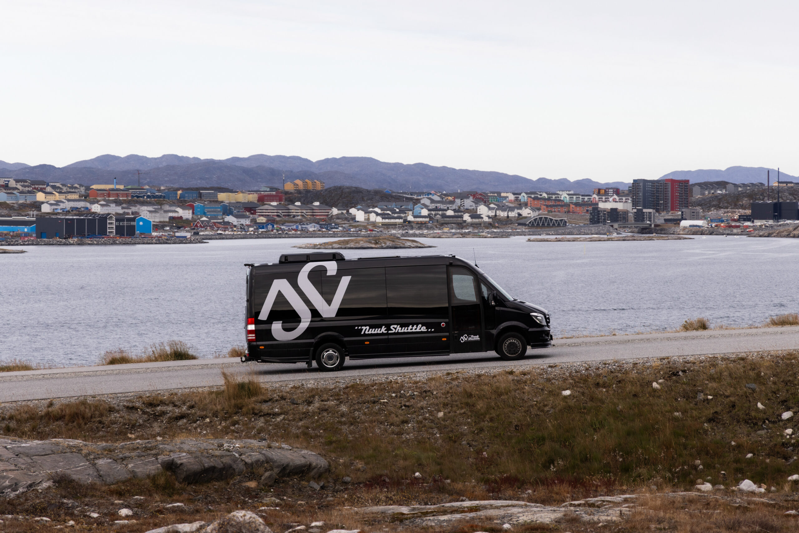 Black shuttle van parked by the water with a town and mountains in the background