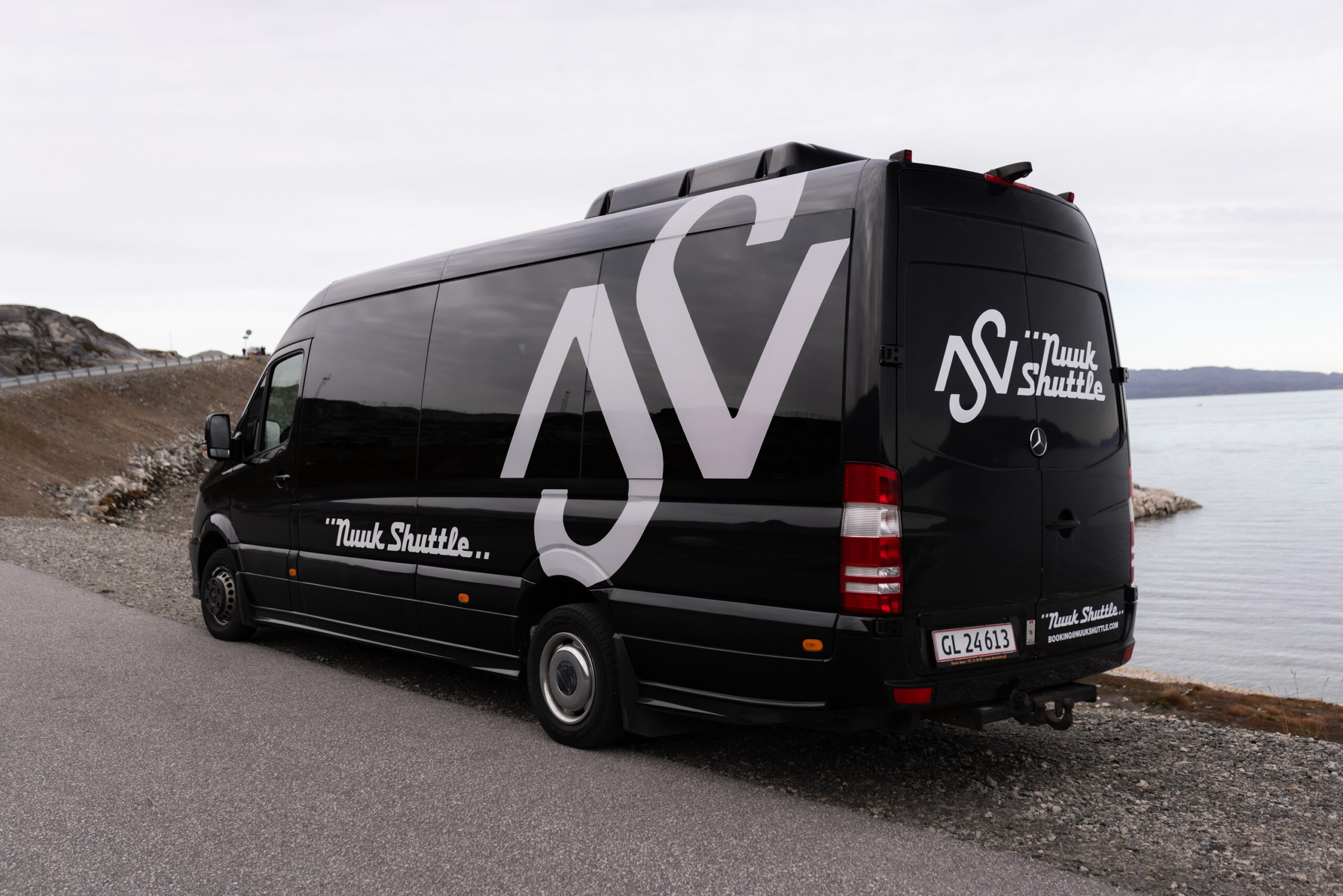 Black shuttle van parked beside a coastal road with water in the background