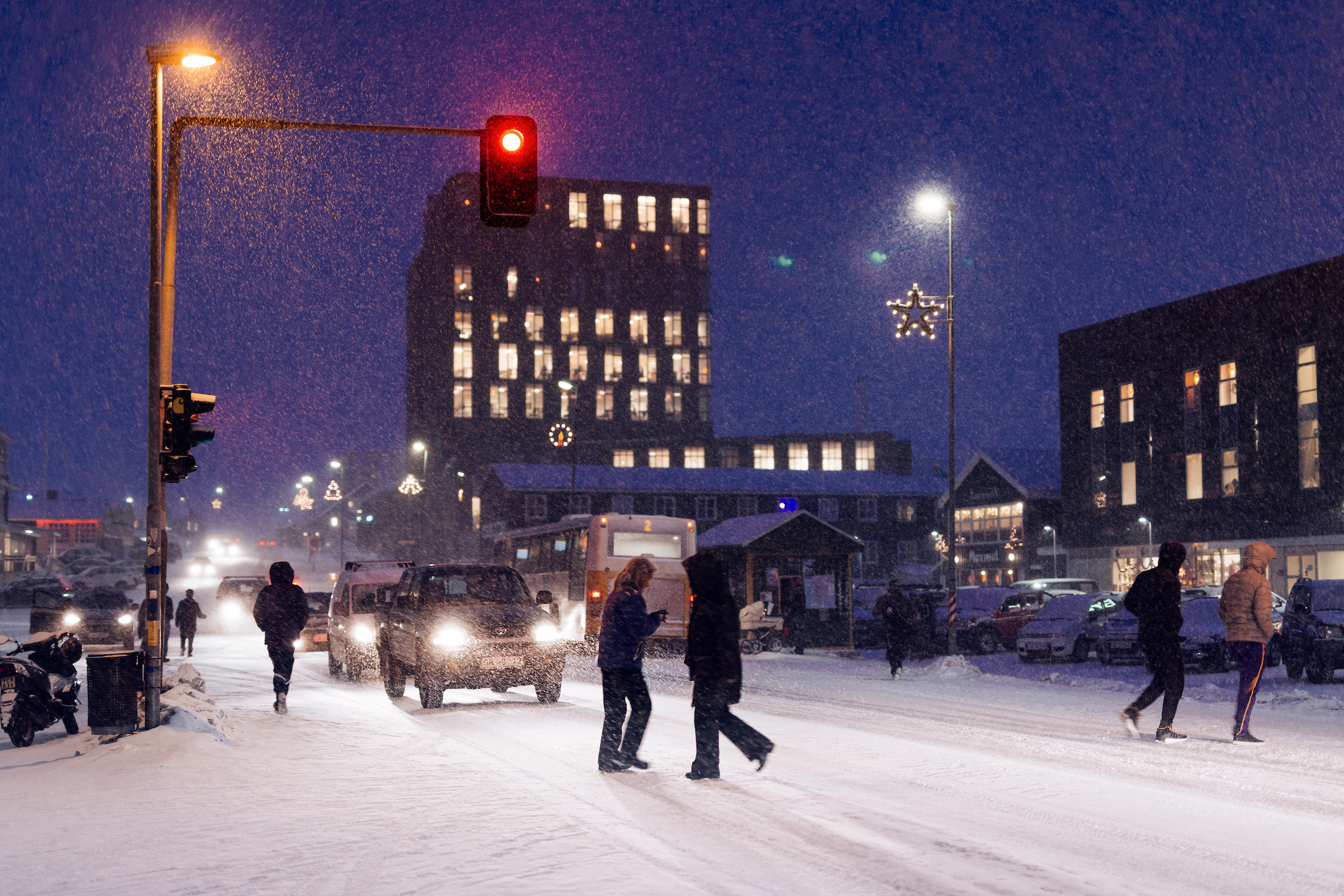Snowy Nuuk city street at dusk with pedestrians, cars, and a red traffic light